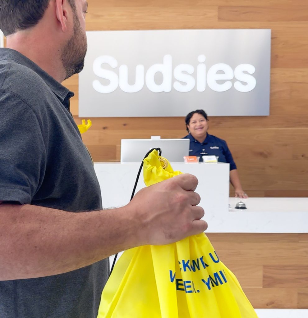 Guest dropping off garments at the Sudsies Boca Raton boutique while a team member welcomes them from behind the counter.