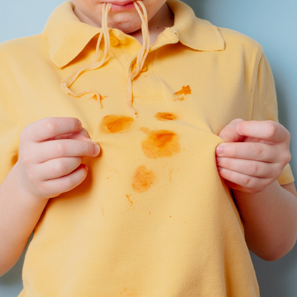 Child holding a yellow shirt stained with spaghetti and tomato sauce while eating noodles.
