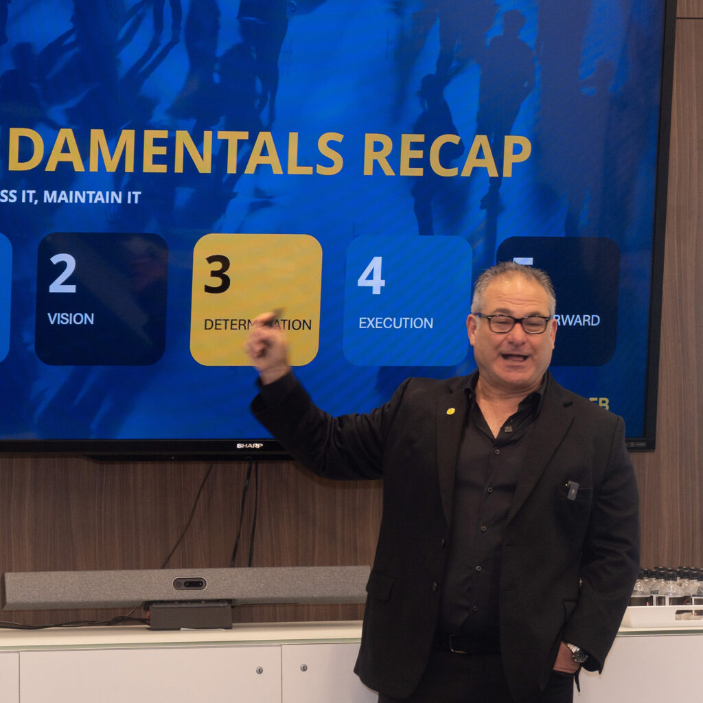 Man in a black suit presenting in front of a large screen that reads “Damentals Recap”