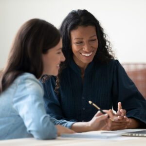 Two women sitting together and smiling while reviewing notes and a laptop during a mentoring session.