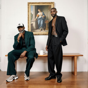 Fashion-forward portrait of a man in relaxed black tailoring standing beside classical artwork in a minimalist interior
